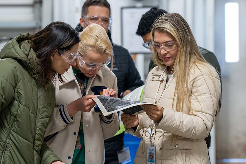 Three women wearing safety goggles are holding an item and examining it closely.