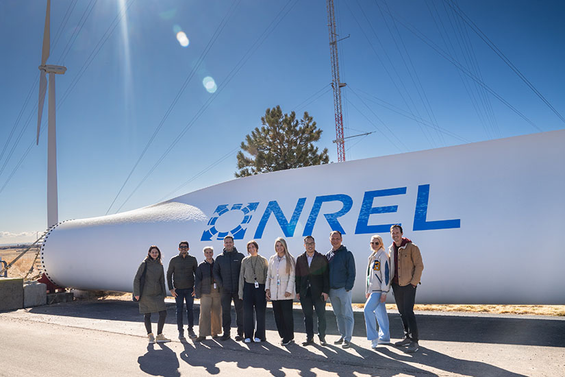 A group of ten people stand smiling for a photo in front of a wind turbine blade with NREL's logo on it