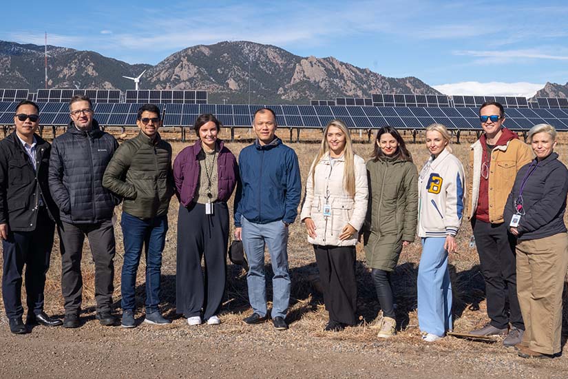 Ten people stand smiling for a photo in front of a solar away and wind turbines. There are mountains in the background.