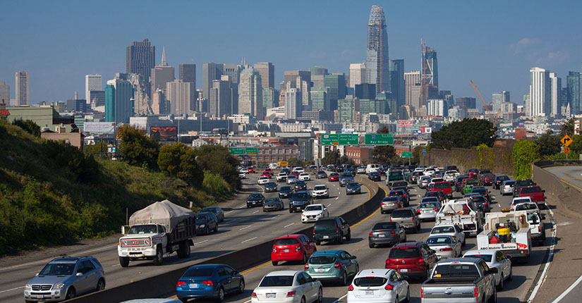 Multiple vehicles on a freeway with a cityscape in the background.
