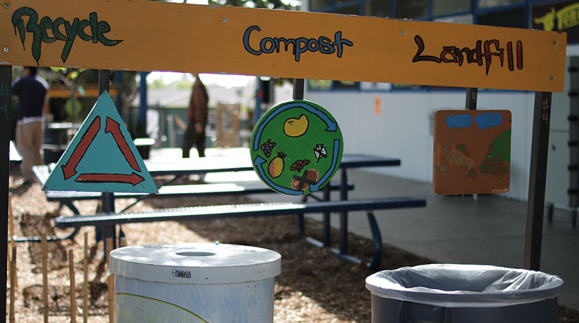 Two bins outside a school with signs over them with the words Recycle - Compost - Landfill. A picnic bench can be seen in the background.
