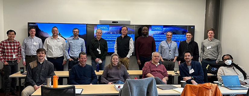 Sixteen researchers in a conference room stand in front of monitors depicting slides showing the NREL logo.