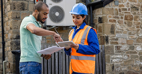 A woman in a construction helmet and safety vest reviews a clipboard with a man.