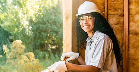 A woman in a construction helmet poses by a window inside an unfinished building.