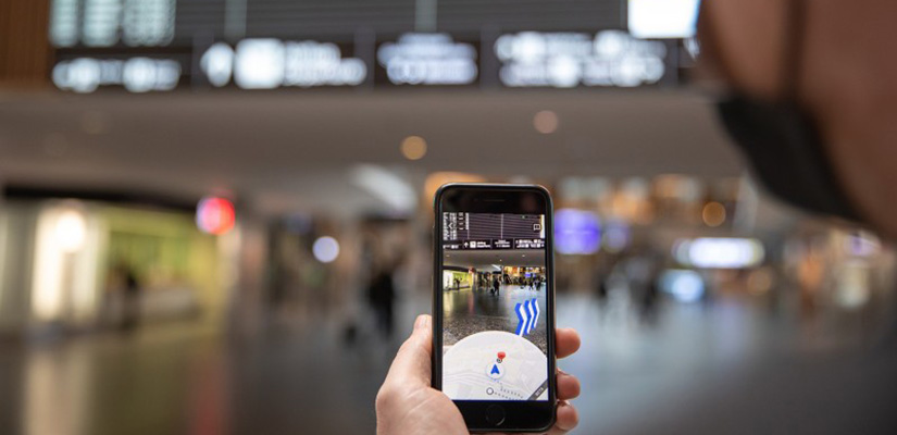 A man holds a smartphone camera up to an airport lobby. On his phone screen, three blue and white arrows point him to the right.