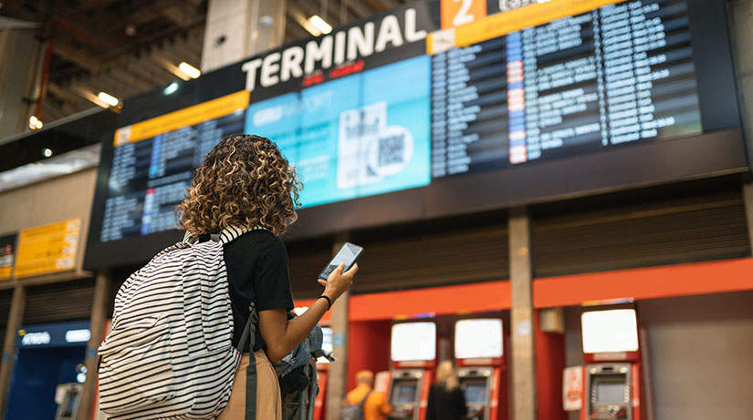 A woman with curly hair faces away from the camera, toward a flight information sign that shows departing planes. She holds up a smartphone to consult it.