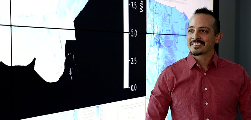 Photo of a person standing and smiling in front of a screen that shows modeled maps.