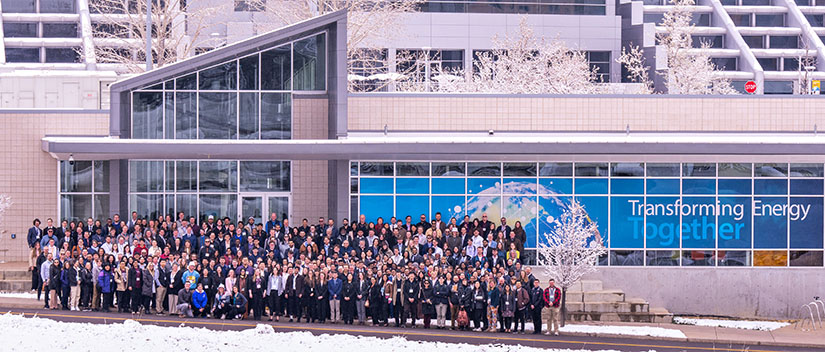 Hundreds of students stand in front of a building with a window cling that reads Transforming Energy Together.