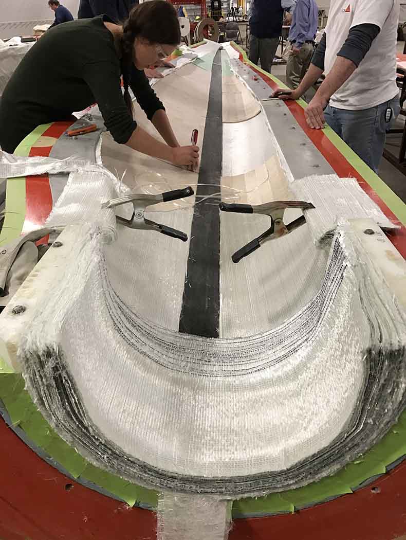 Two people in protective attire standing in a manufacturing facility next to layers of material in a turbine mold.