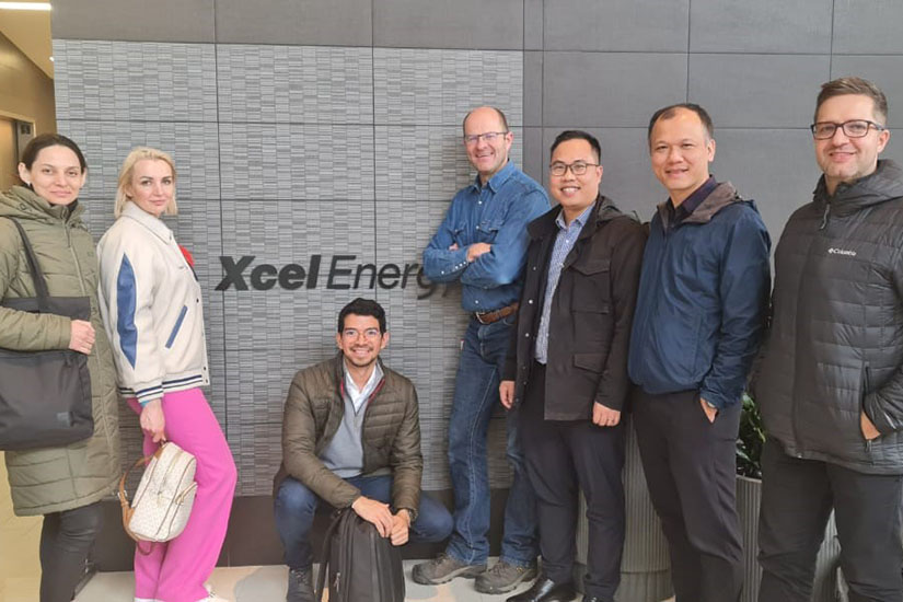 A group of seven individuals standing together and smiling, in front of a wall with a sign for Xcel Energy.