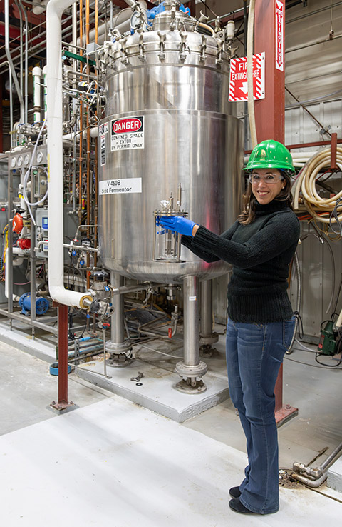 A researcher in a hard hat holds a bioreactor and stands in front of a large bioreactor.