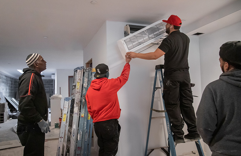 Four men install an HVAC system in a building. One stands on a ladder holding the equipment.