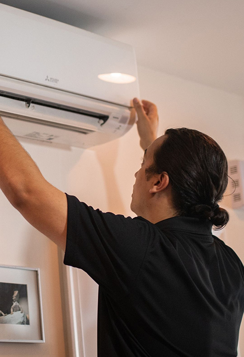 A man in a black shirt fits a rectangle HVAC unit into an apartment wall and ceiling.