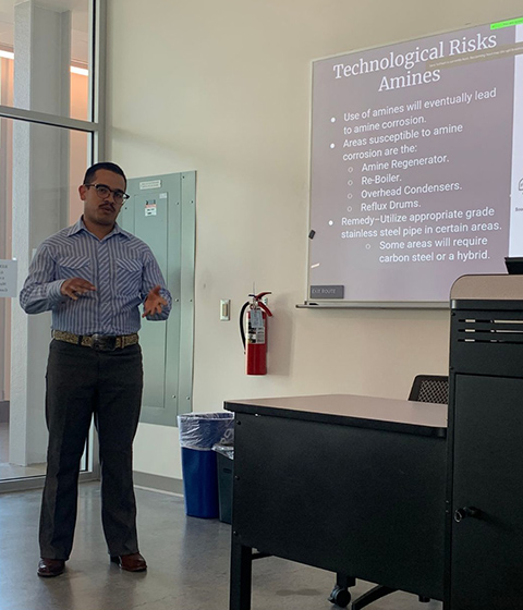 A student stands at the front of a classroom presenting with a slide on a screen behind him.