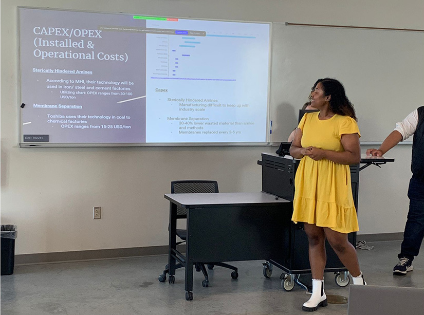 A student in a yellow dress stands at the front of a classroom presenting with a slide on a screen behind her.