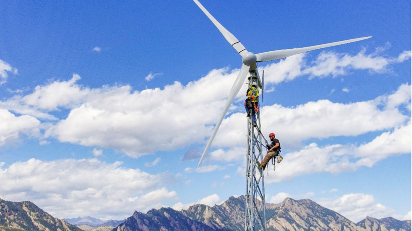 Two people climb a wind turbine with mountains in the background