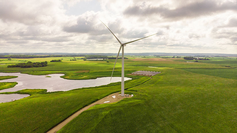 A large wind turbine on a grassy plain near a solar photovoltaic array and a lake.