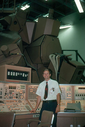 A person stands in from of an old computer model with large metal structures behind him