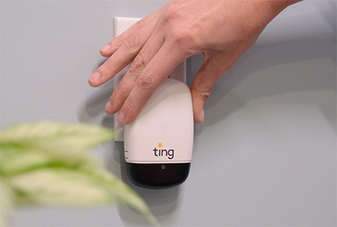 A picture of a small sensor with the name Ting on it plugged into a wall outlet. A hand is just above it after plugging it in. 