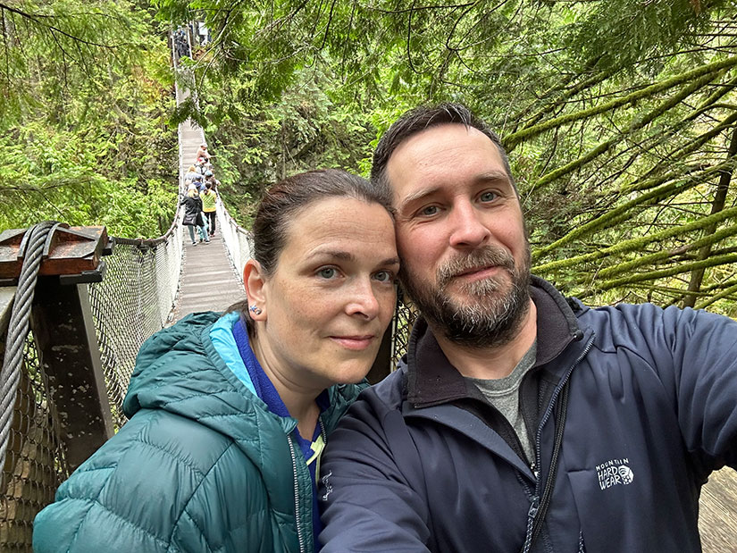 Una selfie de dos personas en un puente colgante con gente caminando sobre el puente detrás de ellos y árboles rodeándolos.
