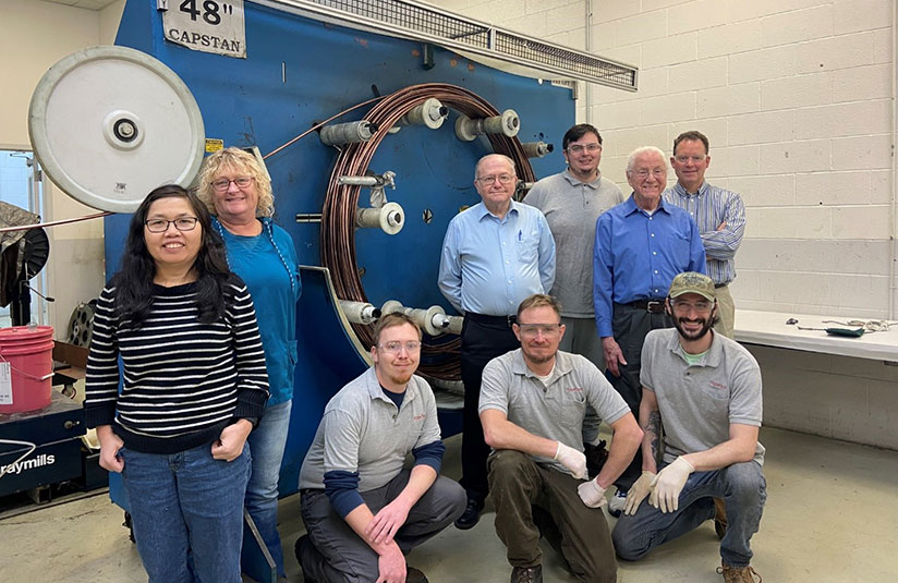 Nine people posing in front of a large coil of wire