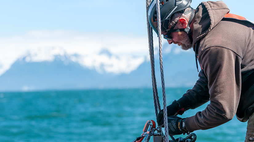 A man pulling on rope on a boat with water and snow-capped mountains in the background