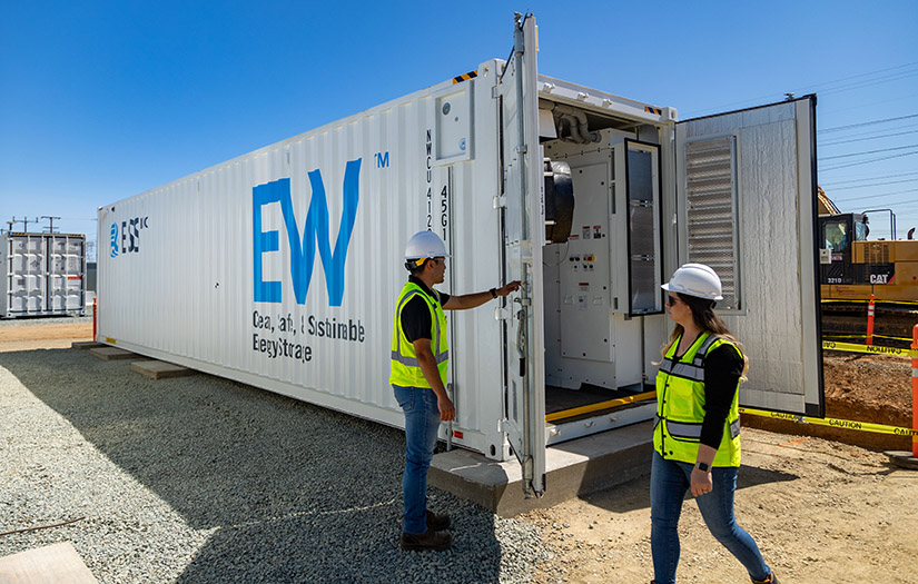 Two people in construction outfits stand near the front of an open storage container with the battery inside.
