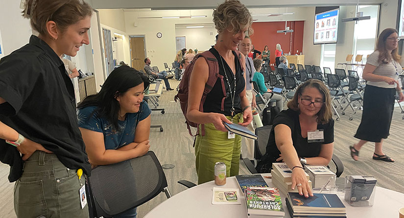 A person sits at a table with books as others stand around the table.