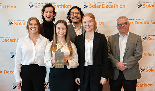 Students and a professor hold a trophy in front of a Solar Decathlon banner at an awards ceremony.