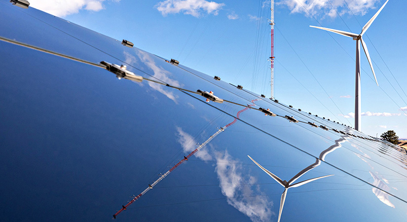 Photo of a solar panel with a wind turbine in the background and blue skies.