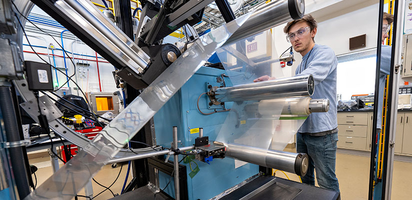 A researcher in a laboratory watching a continuous sheet of material run around multiple metal rolls.