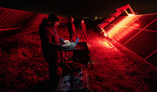 Three people stand near a PV array at night. A bright red light from a scanning instrument lights up the PV modules.