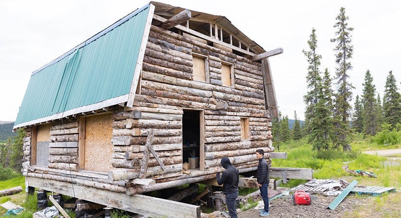 Two individuals stand in front of a log cabin under construction.