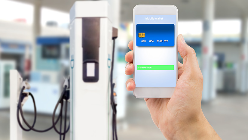 A hand holding a phone with a virtual credit card on the screen and an electric vehicle charging station in the background.