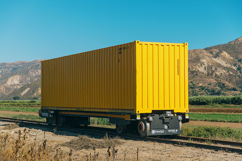 A yellow Parallel Systems autonomous battery-electric rail car against a mountain backdrop.