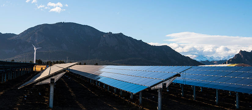 Solar panels in the foreground with mountains and wind turbines in the background.