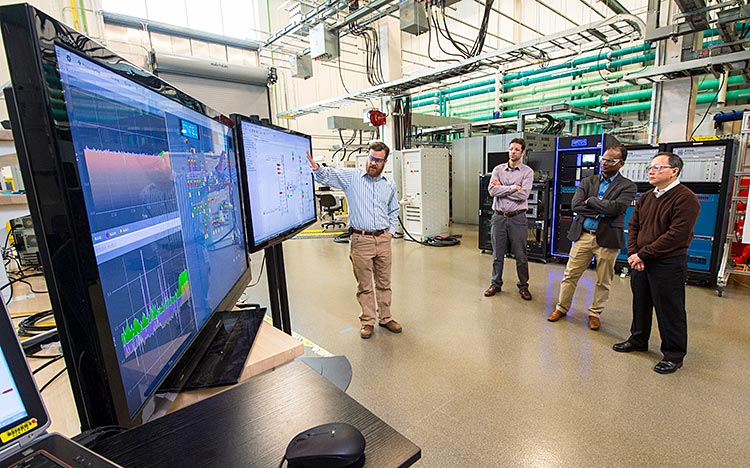 Four people standing in front of large monitors showing microgrid schematics.
