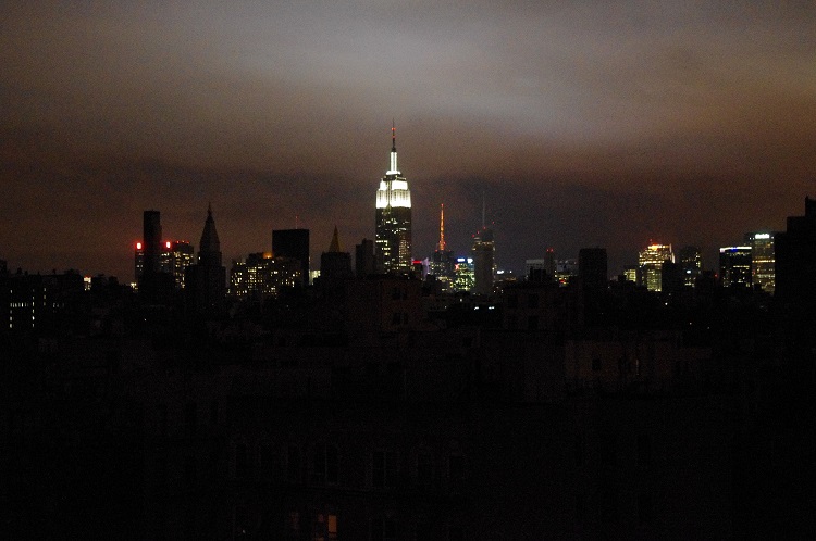 New York City skyline with Midtown and the Empire State Building lights in the background and the darkened East Village and other parts of downtown in the foreground.