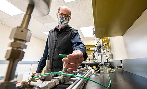 Photo of an NREL researcher, wearing a mask, in a lab with wires and metal casings.
