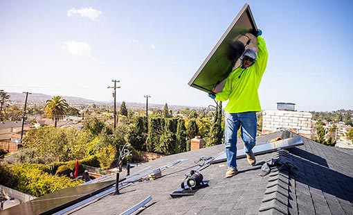 Man wear protective equipment carrying a solar panel on a residential roof.