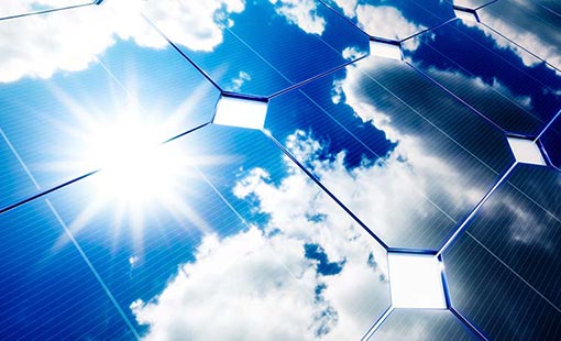 Photo of a close-up view of a solar panel with sun and blue skies in the reflection.