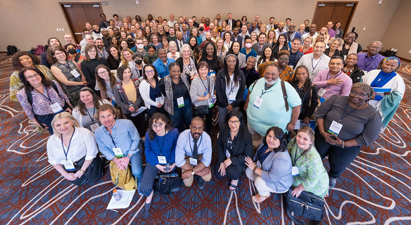 A large group of symposium participants pose with smiles in a hotel conference room.