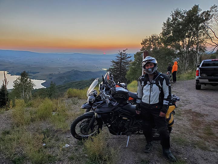 A man with a helmet on standing in front of a motorcycle with a sunset in the background.