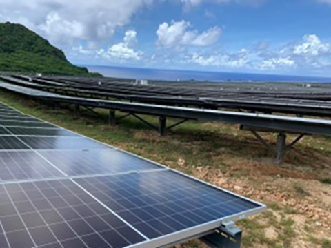 Solar panels in front of a green hill and the ocean.