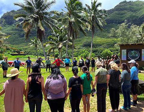 At least 24 people stand hand-in-hand in a circle on green grass beneath palm trees.