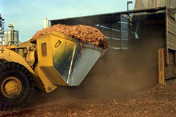 Photo of bulldozer lifting biomass material with factory in background.