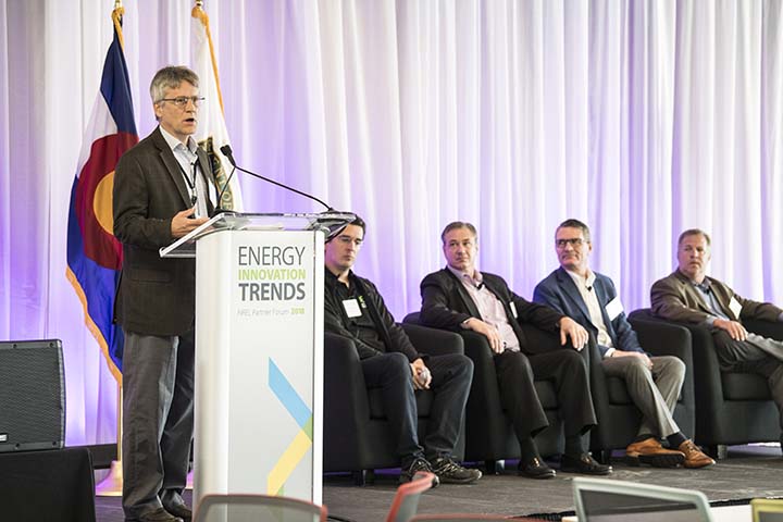 A man speaks at a podium that says Energy Innovation Trends, in front of the Colorado flag. Four other men seated behind him listen to the presentation.