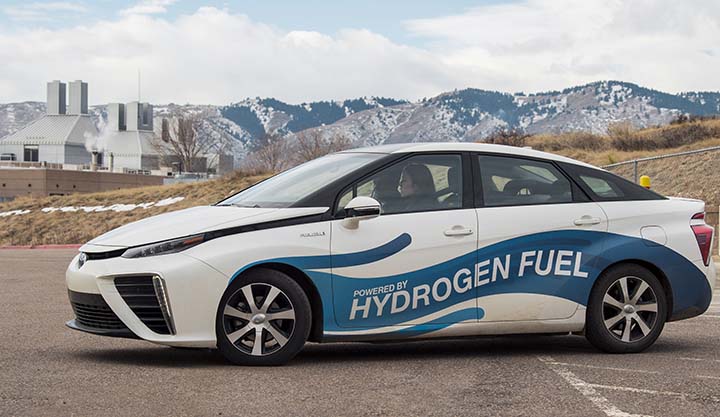 A female driver sits at the wheel of a hydrogen fuel cell vehicle at NREL with mountains and a building in the background. The vehicle has a decal wrap that reads Powered by Hydrogen Fuel.