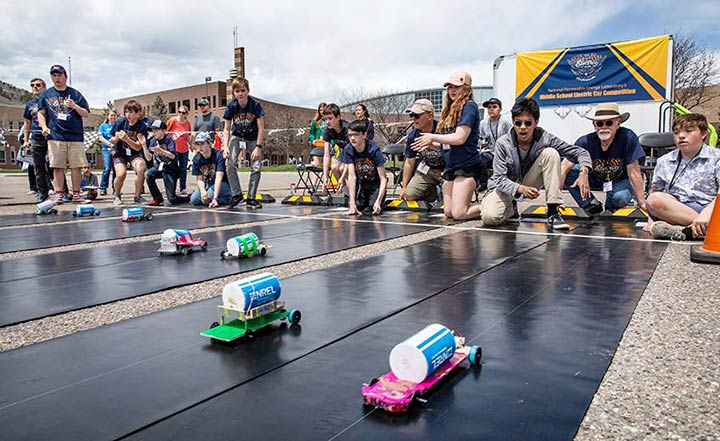 Model cars race down a track as students and adults cheer at a model electric car competition