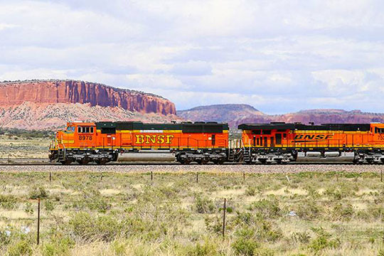 An orange Burlington Northern Santa Fe train races across a colorful desert landscape.